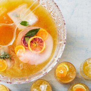 Crystal punch bowl filled with orange punch, ice, mint leaves, and citrus slices, with small glasses of orange slices nearby.